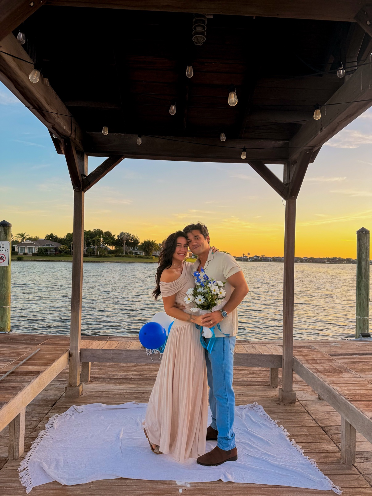 Couple holding a blue and white bouquet together beneath a waterfront pavilion at sunset