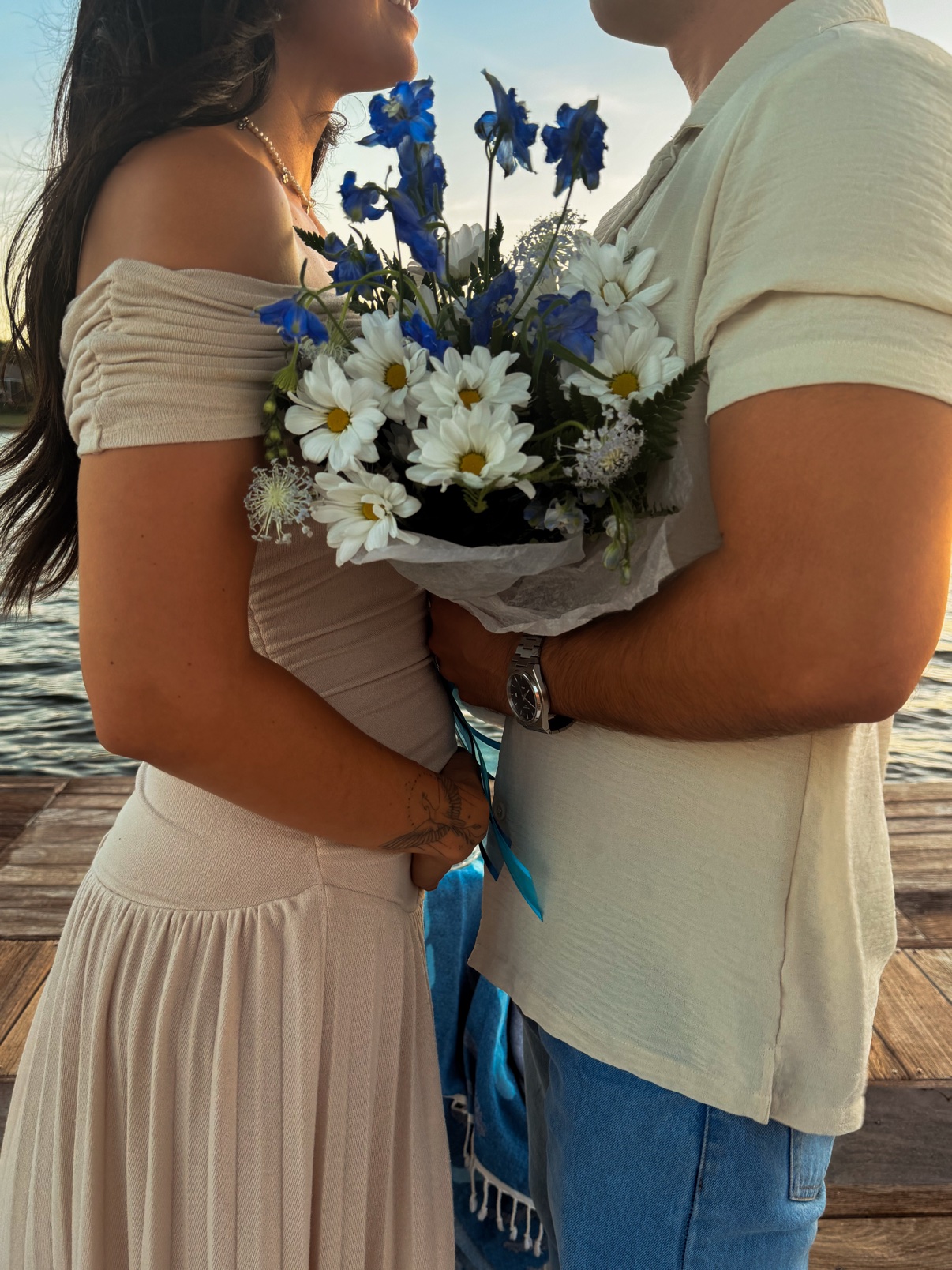 Couple holding a blue and white daisy and delphinium bouquet by the water at sunset