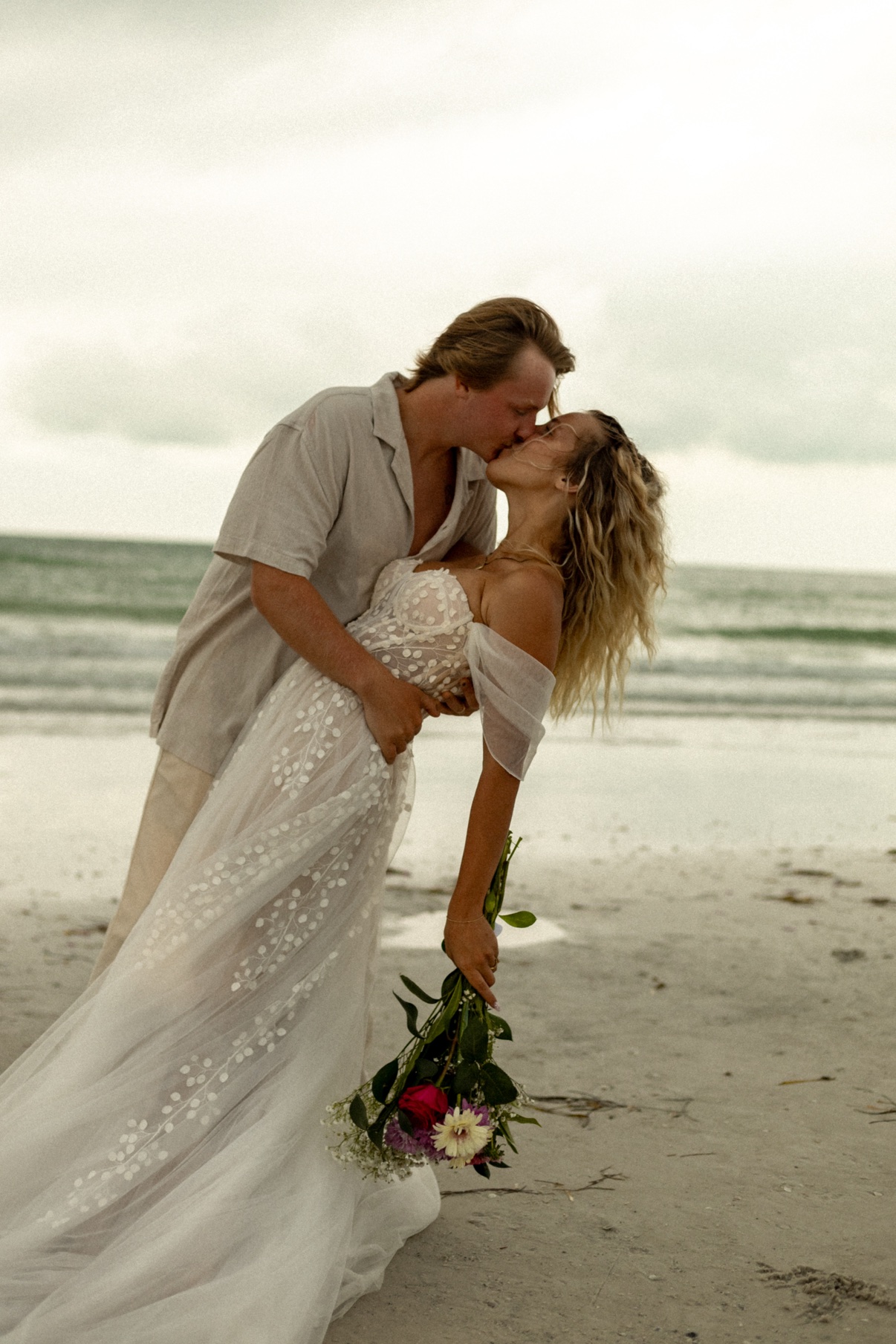Newlyweds kissing on the beach while the bride holds a colorful bouquet
